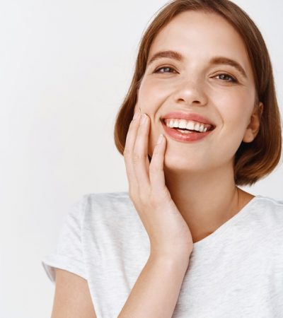 Beauty. Close-up of happy woman with short hair, touching clean fresh facial skin and smiling, showing perfect teeth and face, standing against white background.