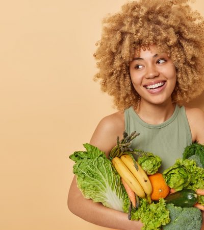 Horizontal shot of beautiful woman carries variety of fresh vegetables and fruits consumes grocery full of vitamins looks away stands against beige background blank space for your promotional content